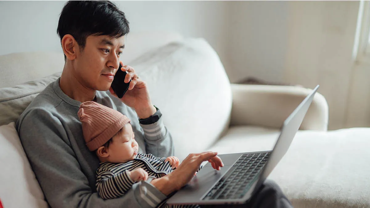 Man sitting on a sofa with a baby on his lap. He&rsquo;s searching the internet and using his mobile phone