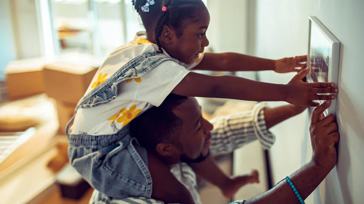 Father and daughter hanging a picture on the wall, the girl is on her father&rsquo;s shoulders